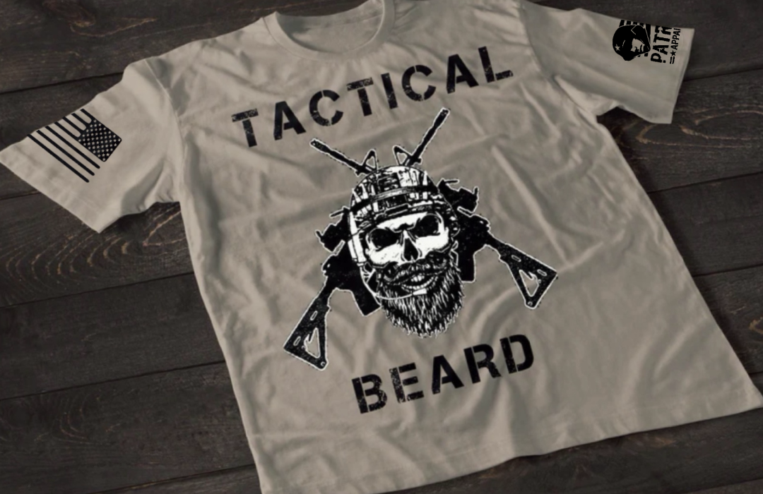 A gray short-sleeve T-shirt laid flat on a dark wooden surface, featuring a bold white graphic of a bearded skull wearing tactical headgear with crossed rifles behind it. The word “TACTICAL” is printed above the design and “BEARD” below. An American flag patch is visible on one sleeve, and a small emblem is printed on the other sleeve.