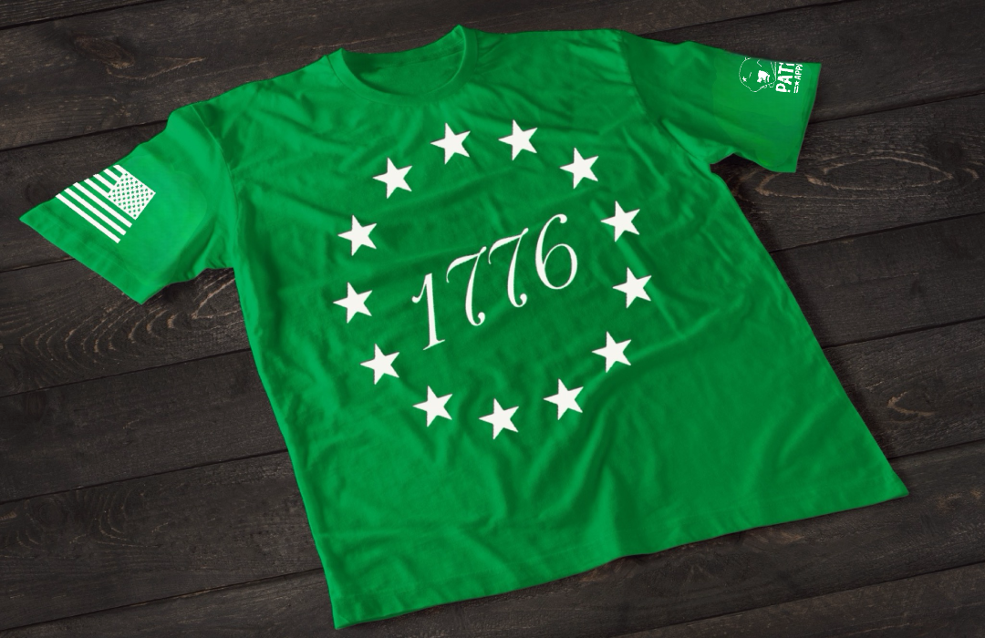 A green short-sleeve T-shirt laid flat on a dark wooden surface, featuring a circular arrangement of thirteen white stars surrounding the number “1776” in the center, inspired by the Betsy Ross flag. An American flag patch is displayed on one sleeve, with a small emblem printed on the other sleeve.
