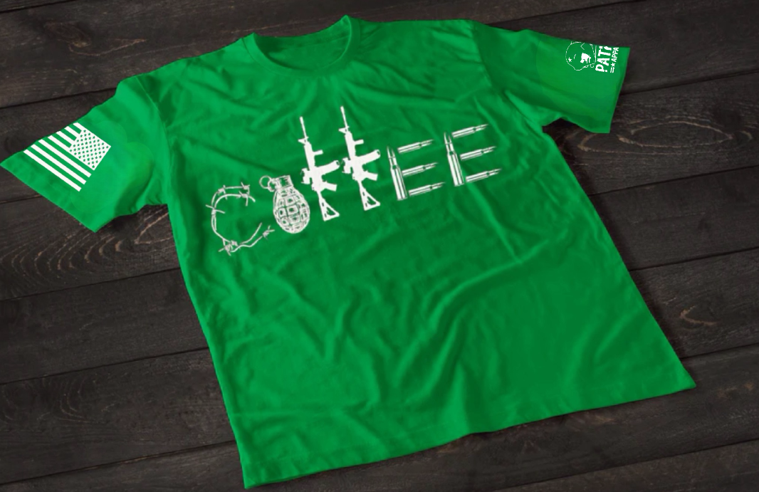 A green short-sleeve T-shirt laid flat on a dark wooden surface, featuring a stylized white graphic spelling “COFFEE,” with each letter formed by different tactical elements such as barbed wire, a grenade, rifles, and ammunition. An American flag patch is visible on one sleeve, and a small emblem is printed on the other sleeve.