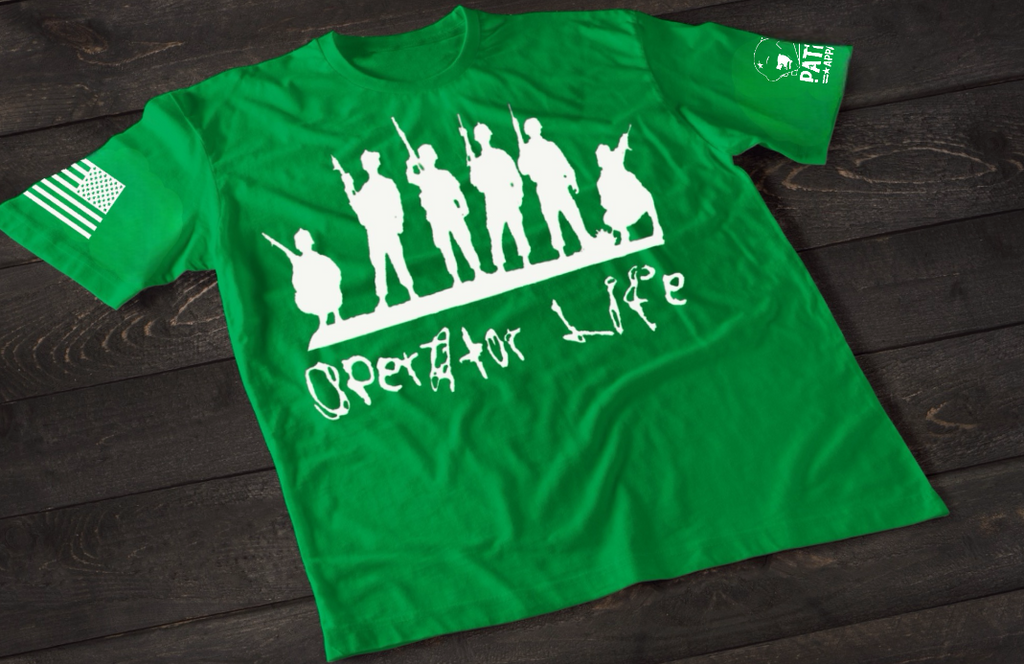 A green short-sleeve T-shirt laid flat on a dark wooden surface, featuring a white graphic of multiple soldier silhouettes holding rifles in different stances. Below the figures is the stylized text “Operator Life.” An American flag patch appears on one sleeve, and a small emblem is printed on the other sleeve.