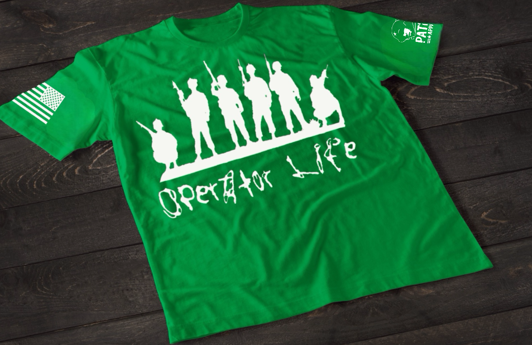 A green short-sleeve T-shirt laid flat on a dark wooden surface, featuring a white graphic of multiple soldier silhouettes holding rifles in different stances. Below the figures is the stylized text “Operator Life.” An American flag patch appears on one sleeve, and a small emblem is printed on the other sleeve.