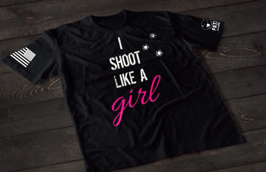 Black women’s I Shoot Like a Girl T-shirt featuring bold white text with pink script lettering and bullet hole graphic accents