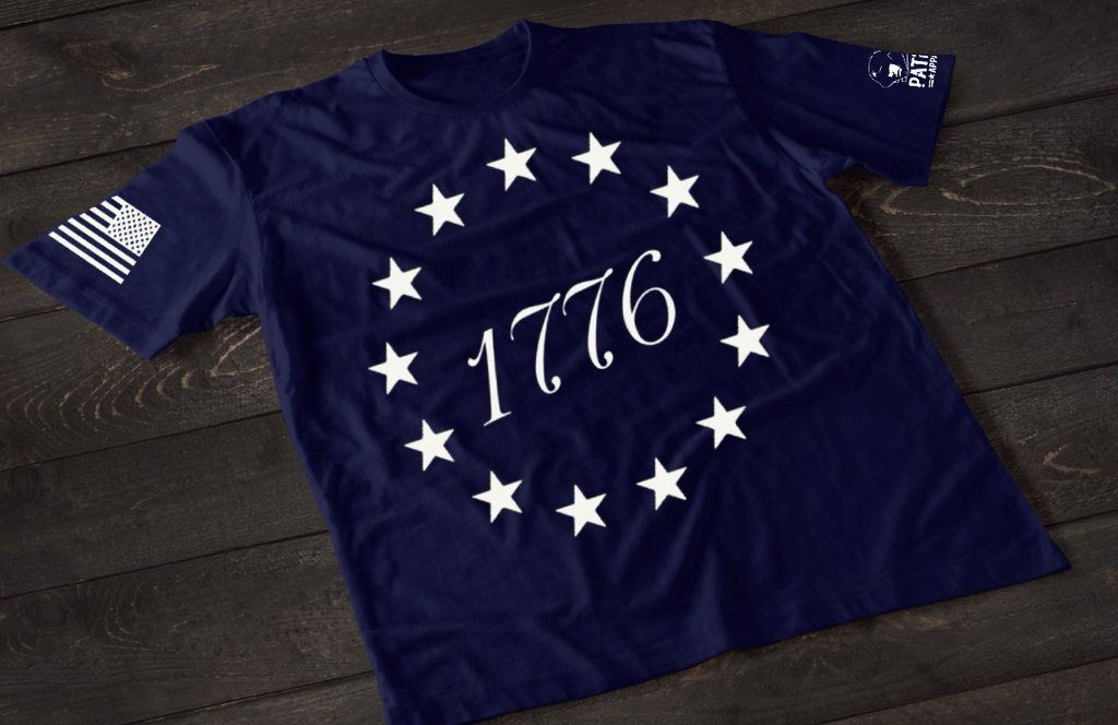 A navy blue short-sleeve T-shirt laid flat on a dark wooden surface, featuring a circular arrangement of thirteen white stars surrounding the number “1776” in the center, inspired by the Betsy Ross flag. An American flag patch is displayed on one sleeve, with a small emblem printed on the other sleeve.