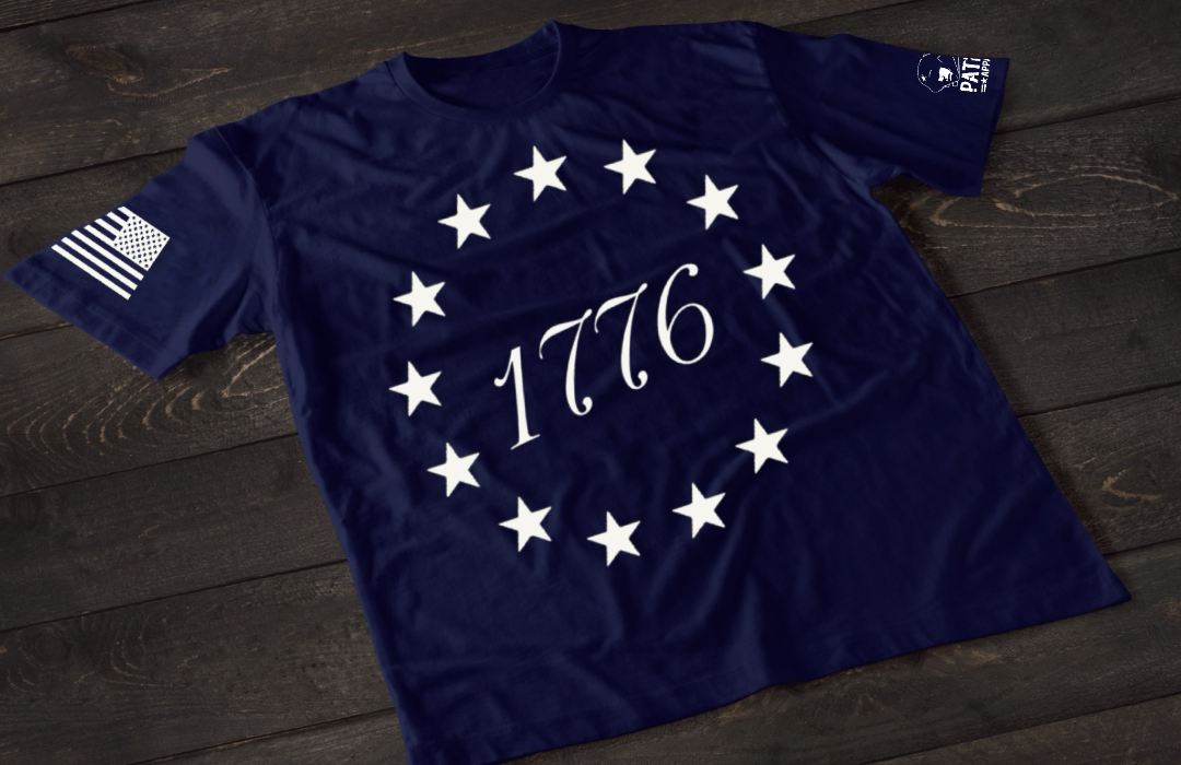 A navy blue short-sleeve T-shirt laid flat on a dark wooden surface, featuring a circular arrangement of thirteen white stars surrounding the number “1776” in the center, inspired by the Betsy Ross flag. An American flag patch is displayed on one sleeve, with a small emblem printed on the other sleeve.