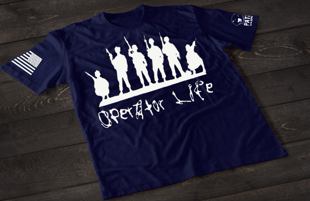 A navy blue short-sleeve T-shirt laid flat on a dark wooden surface, featuring a white graphic of multiple soldier silhouettes holding rifles in different stances. Below the figures is the stylized text “Operator Life.” An American flag patch appears on one sleeve, and a small emblem is printed on the other sleeve.
