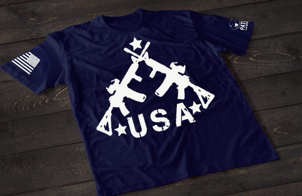 A Navy Blue short-sleeve T-shirt laid flat on a dark wooden surface, featuring a bold white graphic of three crossed rifles above the word “USA” with stars. An American flag patch appears on one sleeve, and a small circular emblem is printed on the other sleeve.