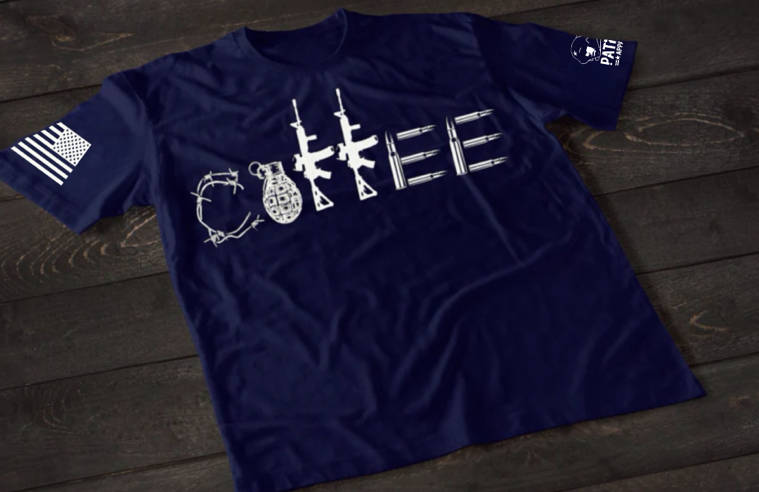 A navy blue short-sleeve T-shirt laid flat on a dark wooden surface, featuring a stylized white graphic spelling “COFFEE,” with each letter formed by different tactical elements such as barbed wire, a grenade, rifles, and ammunition. An American flag patch is visible on one sleeve, and a small emblem is printed on the other sleeve.