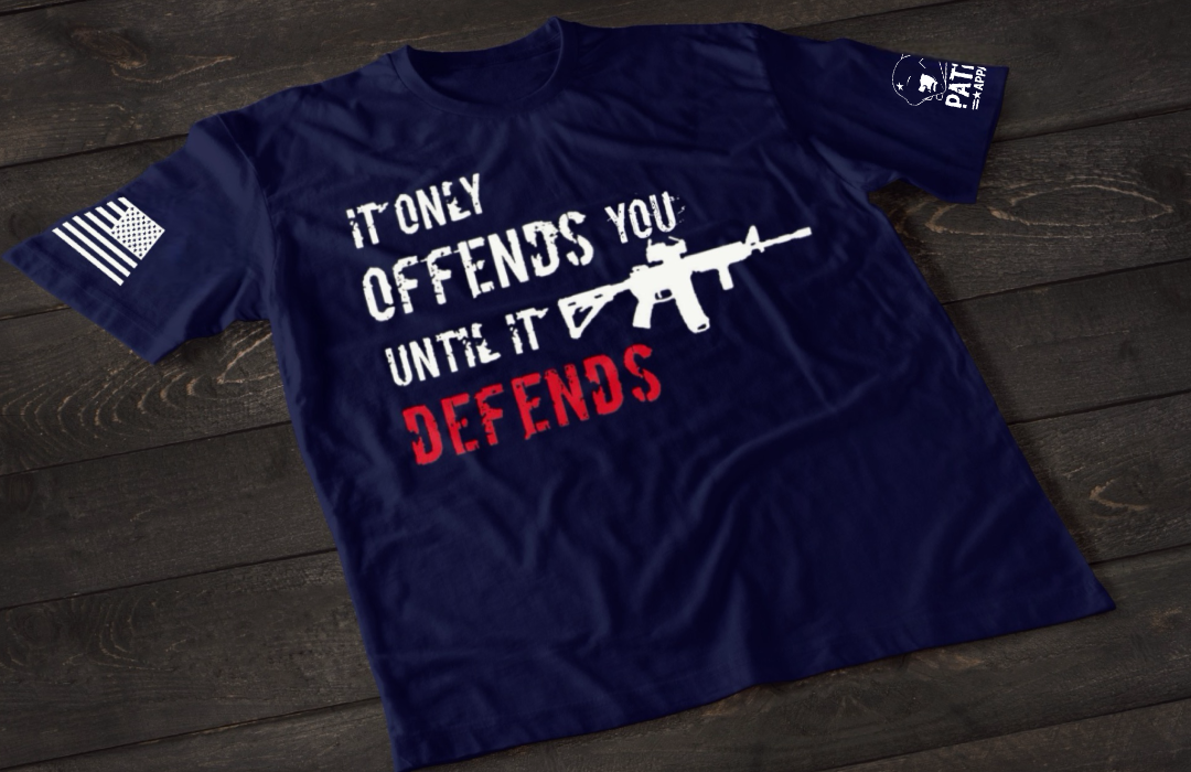 A navy blue short-sleeve T-shirt laid flat on a dark wooden surface, featuring bold distressed text that reads “IT ONLY OFFENDS YOU UNTIL IT DEFENDS,” with the word “DEFENDS” in red. A white rifle graphic is incorporated into the design. An American flag patch is visible on one sleeve, and a small emblem is printed on the other sleeve.