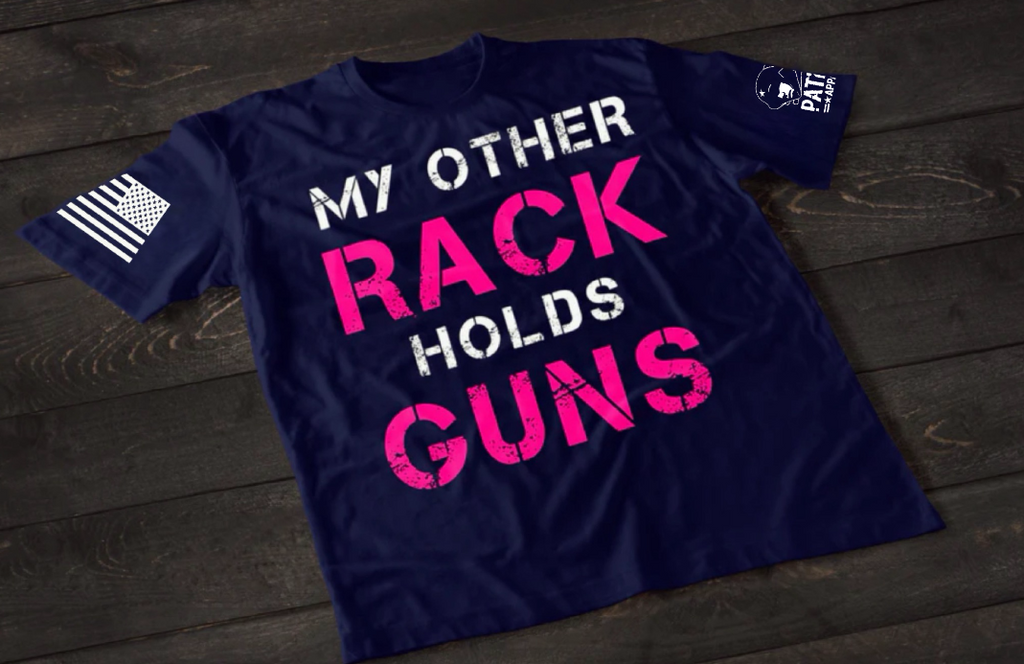 A navy blue short-sleeve T-shirt laid flat on a dark wooden surface, featuring large distressed text that reads “MY OTHER RACK HOLDS GUNS,” with “RACK” and “GUNS” in bright pink and the rest in white. An American flag patch is visible on one sleeve, and a small emblem is printed on the other sleeve.