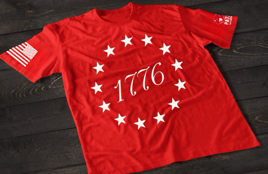 A red short-sleeve T-shirt laid flat on a dark wooden surface, featuring a circular arrangement of thirteen white stars surrounding the number “1776” in the center, inspired by the Betsy Ross flag. An American flag patch is displayed on one sleeve, with a small emblem printed on the other sleeve.
