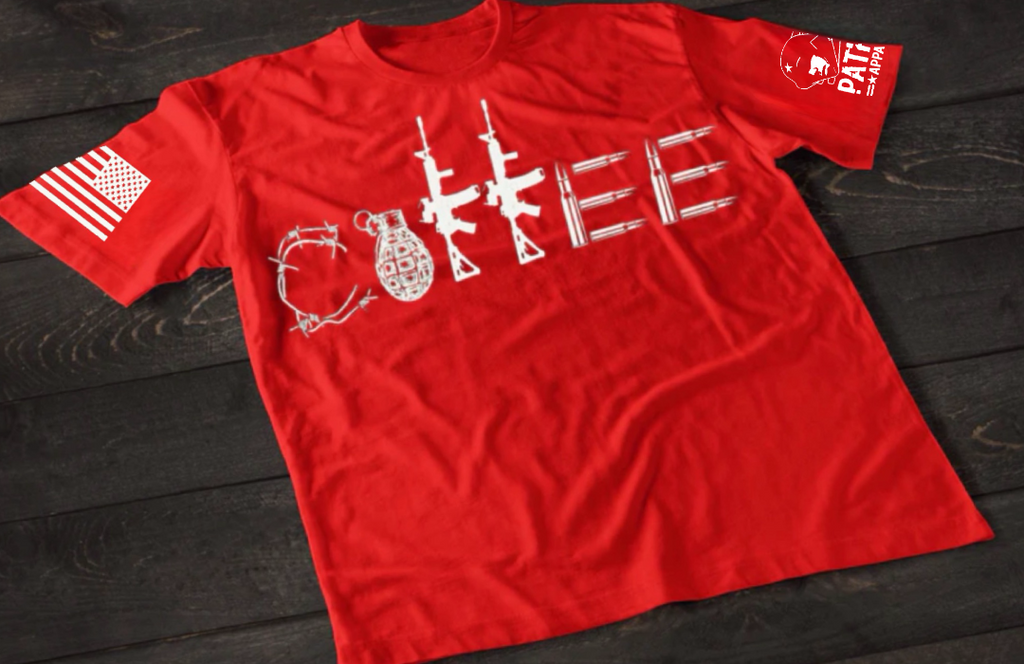 A red short-sleeve T-shirt laid flat on a dark wooden surface, featuring a stylized white graphic spelling “COFFEE,” with each letter formed by different tactical elements such as barbed wire, a grenade, rifles, and ammunition. An American flag patch is visible on one sleeve, and a small emblem is printed on the other sleeve.