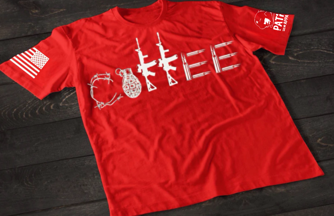 A red short-sleeve T-shirt laid flat on a dark wooden surface, featuring a stylized white graphic spelling “COFFEE,” with each letter formed by different tactical elements such as barbed wire, a grenade, rifles, and ammunition. An American flag patch is visible on one sleeve, and a small emblem is printed on the other sleeve.