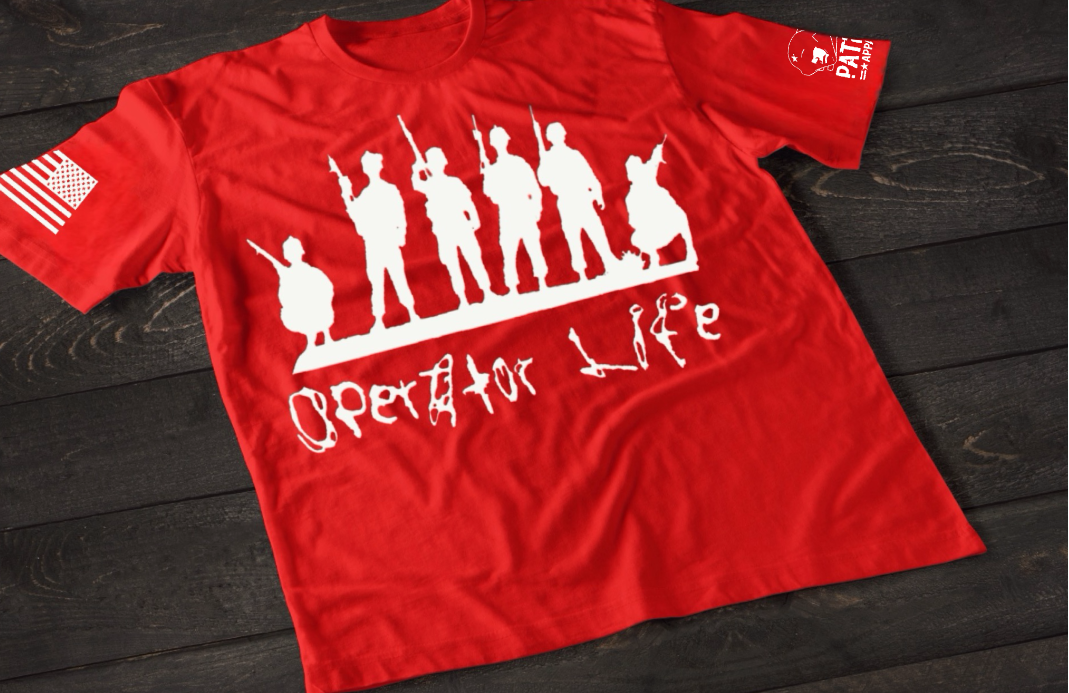 A red short-sleeve T-shirt laid flat on a dark wooden surface, featuring a white graphic of multiple soldier silhouettes holding rifles in different stances. Below the figures is the stylized text “Operator Life.” An American flag patch appears on one sleeve, and a small emblem is printed on the other sleeve.