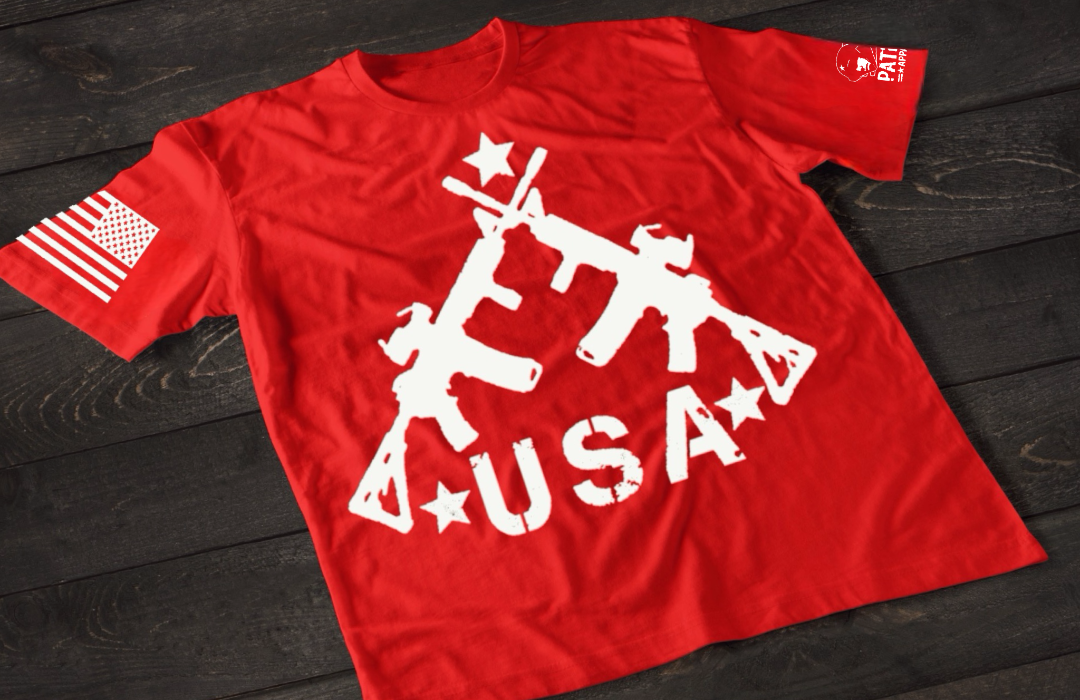 A red short-sleeve T-shirt laid flat on a dark wooden surface, featuring a bold white graphic of three crossed rifles above the word “USA” with stars. An American flag patch appears on one sleeve, and a small circular emblem is printed on the other sleeve.