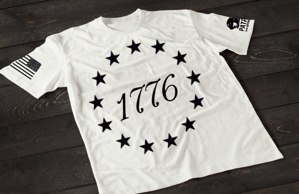 A white short-sleeve T-shirt laid flat on a dark wooden surface, featuring a circular arrangement of thirteen white stars surrounding the number “1776” in the center, inspired by the Betsy Ross flag. An American flag patch is displayed on one sleeve, with a small emblem printed on the other sleeve.