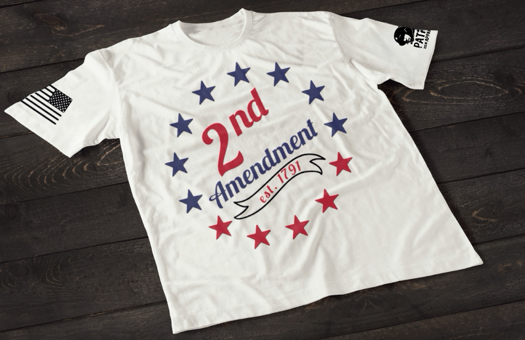 A white short-sleeve T-shirt laid flat on a dark wooden surface, featuring a circular arrangement of red and blue stars surrounding the text “2nd Amendment” in bold lettering, with “est. 1791” displayed on a ribbon below. An American flag patch appears on one sleeve, and a small emblem is printed on the other sleeve.