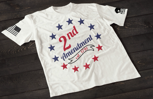 A white short-sleeve T-shirt laid flat on a dark wooden surface, featuring a circular arrangement of red and blue stars surrounding the text “2nd Amendment” in bold lettering, with “est. 1791” displayed on a ribbon below. An American flag patch appears on one sleeve, and a small emblem is printed on the other sleeve.
