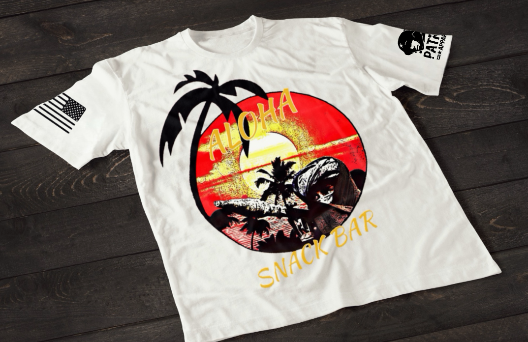 A white short-sleeve T-shirt laid flat on a dark wooden surface, featuring a colorful circular graphic of a tropical beach scene with palm trees, ocean waves, and a sunset in red and yellow tones. The word “ALOHA” appears above the design, with “SNACK BAR” printed below in a stylized font. An American flag patch is visible on one sleeve, and a small emblem is printed on the other sleeve.