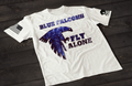 A white short-sleeve T-shirt laid flat on a dark wooden surface, featuring a large distressed-style blue falcon graphic in flight. Above the image is the text “BLUE FALCONS,” with “FLY ALONE” printed below. An American flag patch appears on one sleeve, and a small emblem is printed on the other sleeve.
