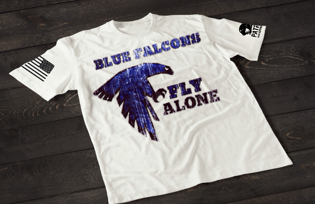 A white short-sleeve T-shirt laid flat on a dark wooden surface, featuring a large distressed-style blue falcon graphic in flight. Above the image is the text “BLUE FALCONS,” with “FLY ALONE” printed below. An American flag patch appears on one sleeve, and a small emblem is printed on the other sleeve.