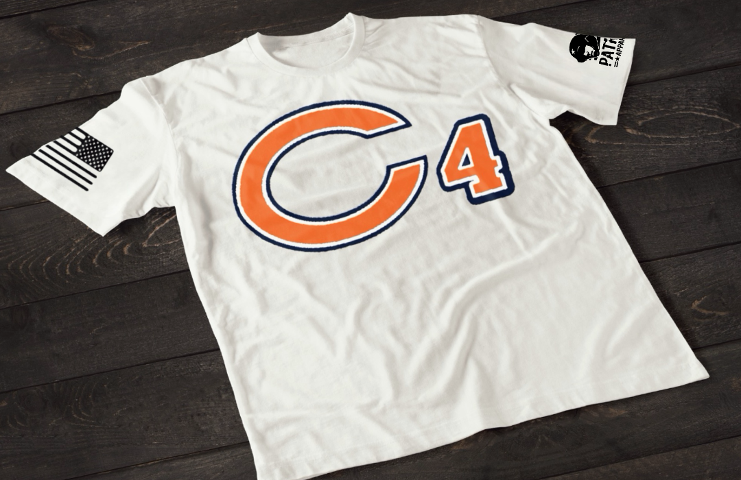 A white short-sleeve T-shirt laid flat on a dark wooden surface, featuring a large orange letter “C” outlined in navy blue with the number “4” beside it in matching colors. An American flag patch is visible on one sleeve, and a small emblem is printed on the other sleeve.