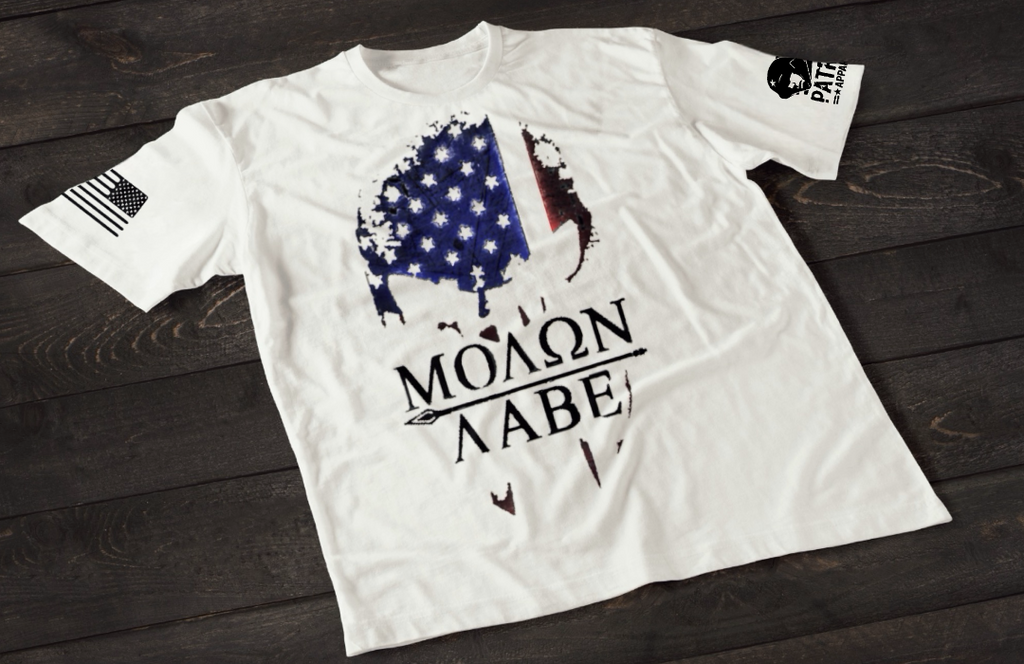 A white short-sleeve T-shirt laid flat on a dark wooden surface, featuring a stylized skull graphic filled with a distressed American flag pattern in blue and red tones. Below the image is the text “MOLON LABE” with a spear running through the lettering. An American flag patch is visible on one sleeve, and a small emblem is printed on the other sleeve.