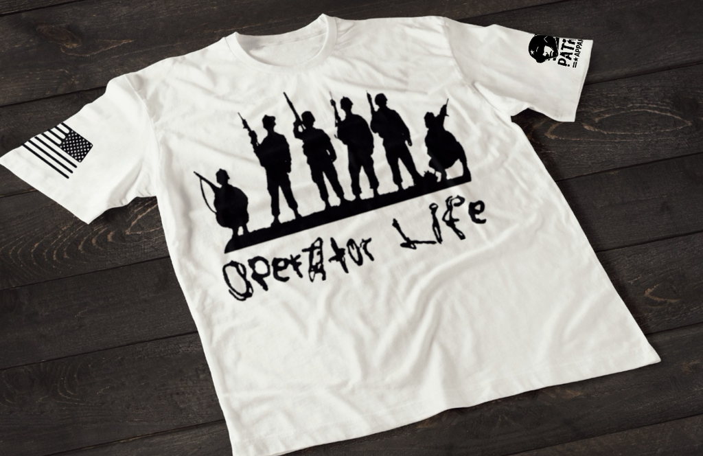 A white short-sleeve T-shirt laid flat on a dark wooden surface, featuring a white graphic of multiple soldier silhouettes holding rifles in different stances. Below the figures is the stylized text “Operator Life.” An American flag patch appears on one sleeve, and a small emblem is printed on the other sleeve.