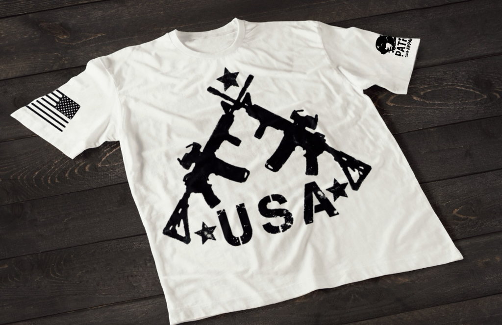 A white short-sleeve T-shirt laid flat on a dark wooden surface, featuring a bold white graphic of three crossed rifles above the word “USA” with stars. An American flag patch appears on one sleeve, and a small circular emblem is printed on the other sleeve.