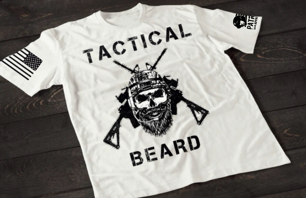 White short-sleeve T-shirt laid flat on a dark wooden surface, featuring a bold white graphic of a bearded skull wearing tactical headgear with crossed rifles behind it. The word “TACTICAL” is printed above the design and “BEARD” below. An American flag patch is visible on one sleeve, and a small emblem is printed on the other sleeve.