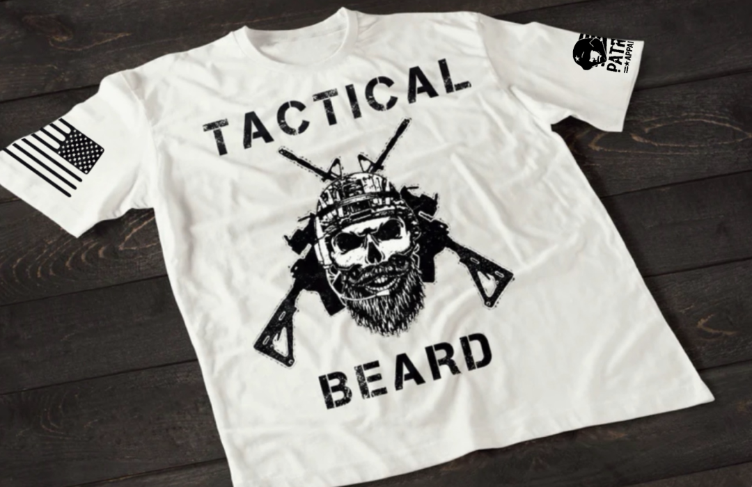 White short-sleeve T-shirt laid flat on a dark wooden surface, featuring a bold white graphic of a bearded skull wearing tactical headgear with crossed rifles behind it. The word “TACTICAL” is printed above the design and “BEARD” below. An American flag patch is visible on one sleeve, and a small emblem is printed on the other sleeve.